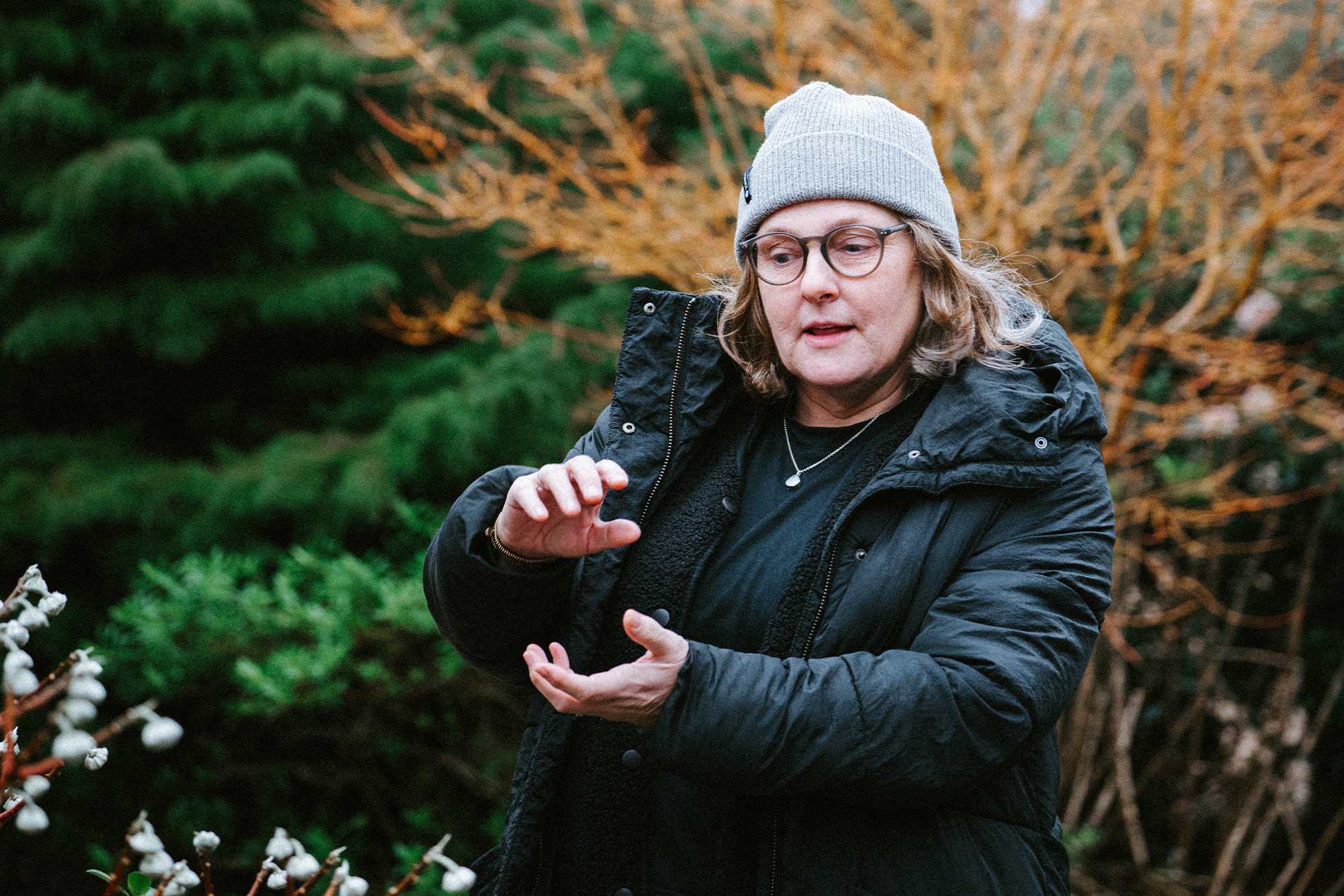 Kate, a middle-aged woman in a grey hat and thick coat stands against a backdrop of trees and plants demonstrating something with her hands