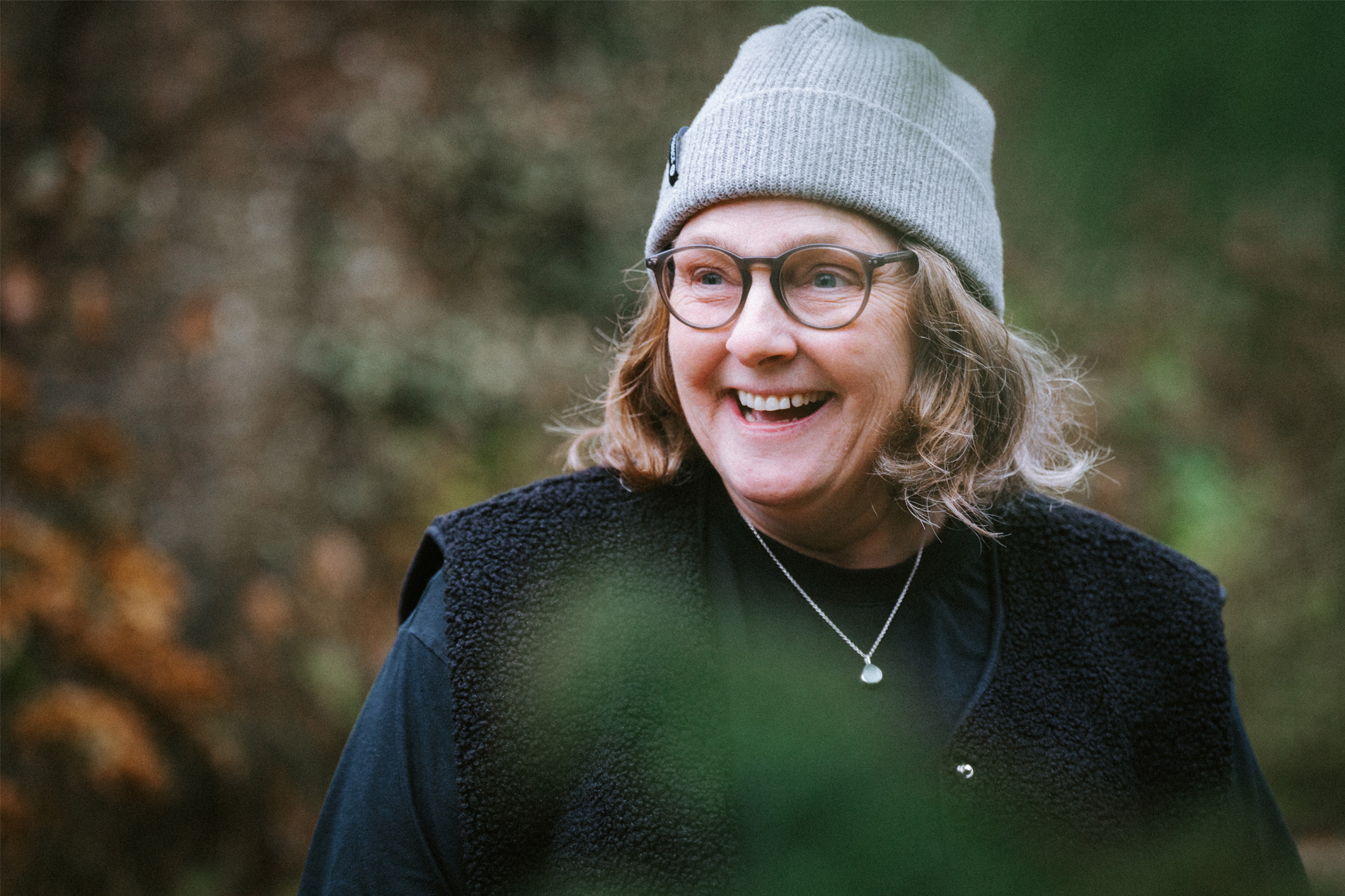 Close-up headshot of Kate, a middle-ages woman wearing a grey hat, smiling against a garden background
