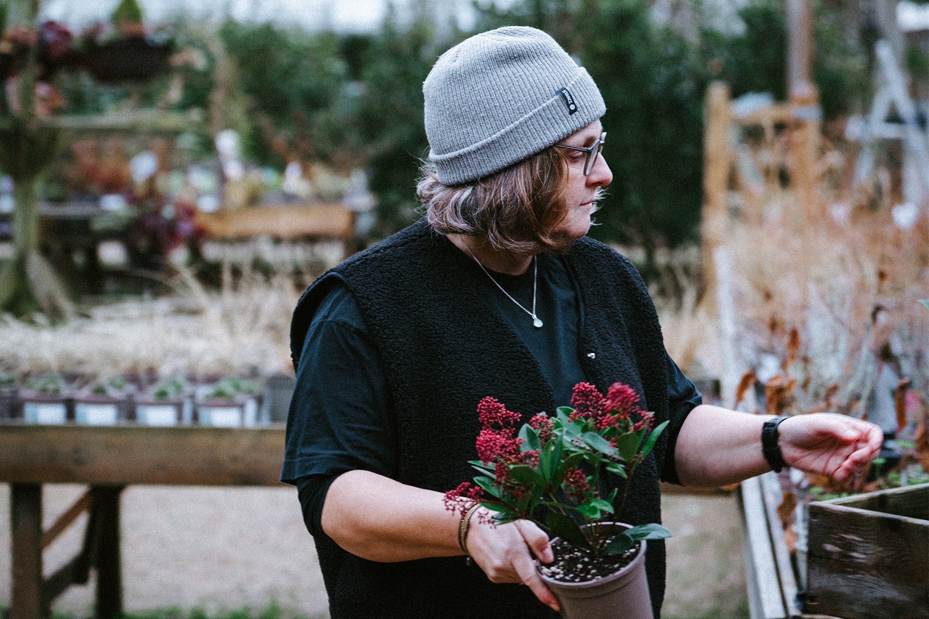 Kate, a middle-aged woman in a grey hat in an outdoor plant nursery holding a red flowering potted plant