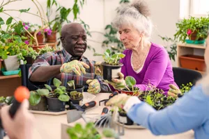 An older black man and white, grey haired woman sit at a table of seedling pots, wearing gardening gloves and appearing to be potting up plants An older black man and white, grey haired woman sit at a table of seedling pots, wearing gardening gloves and appearing to be potting up plants