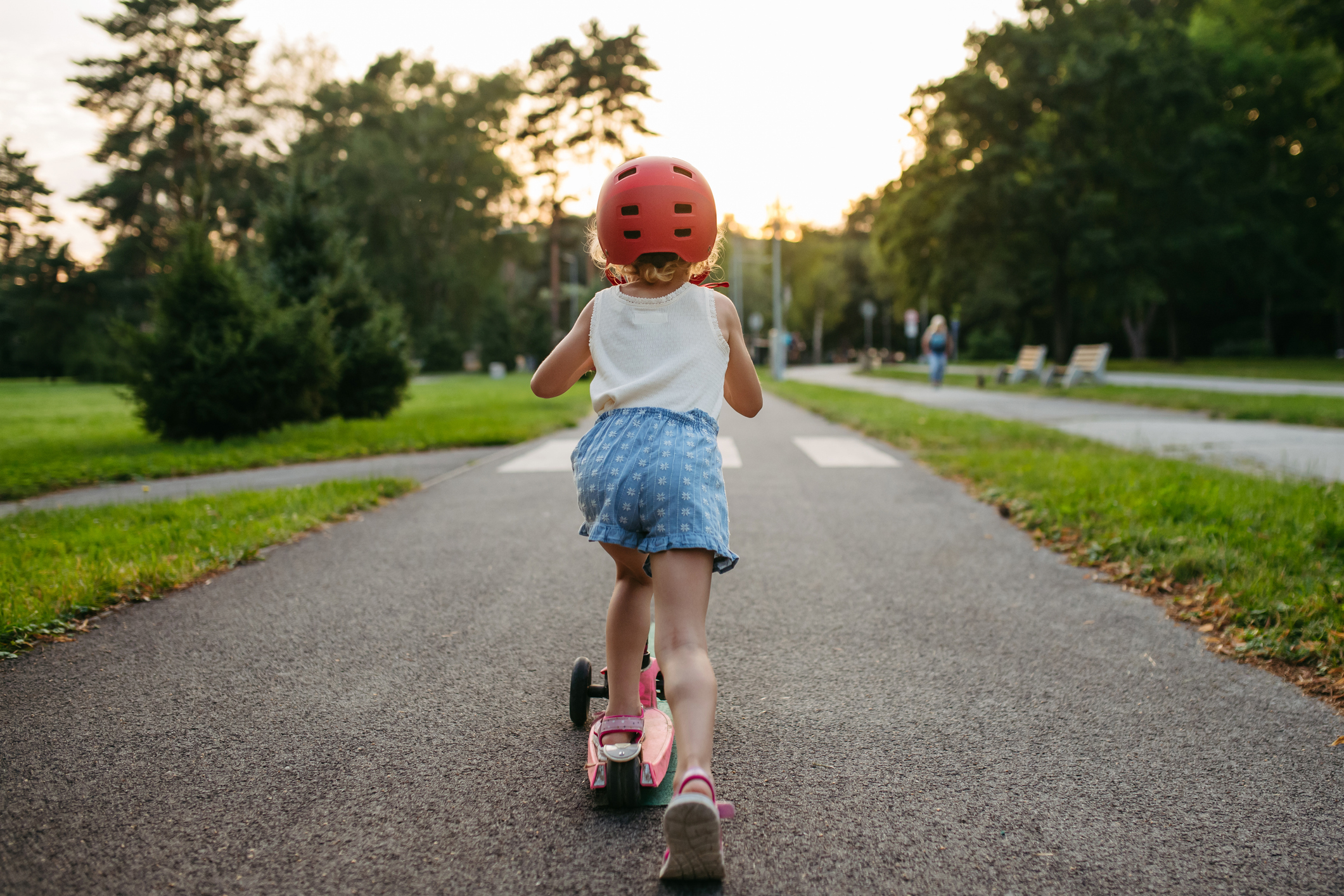 Rear view of cute girl with helmet riding scooter on bike path in the city during sunset.