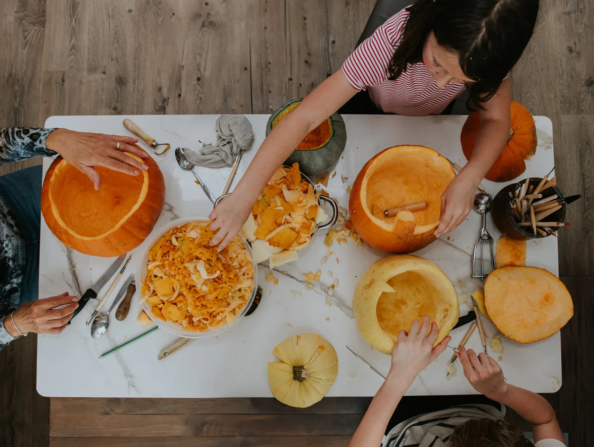 Top down image of halloween fun - Pumpkins are spread across a marble table top, with family members sitting around the table carving pumpkins and making Jack O' Lanterns A family make Jack O' Lanterns for Hallowe'en. They sit around a white marble table and concentrate on carving faces into pumpkins. A messy but fun activity. The pumpkins are completely hollowed out. © Getty / Catherine Falls Commercial