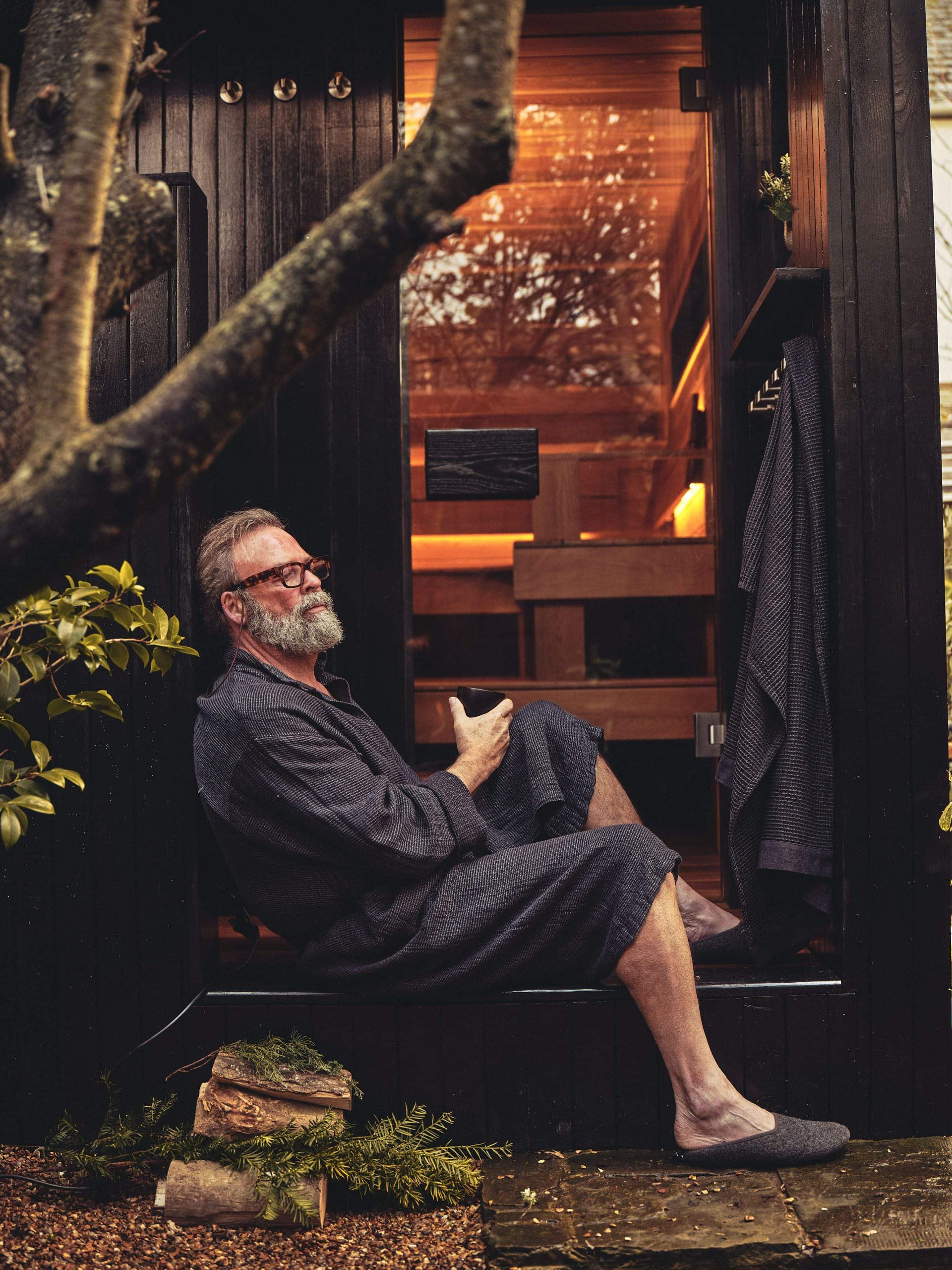Man with grey hair and beard wearing glasses and a dark grey dressing gown and slippers sits on the outside steps of a glass door sauna hut, glowing warm light inside
