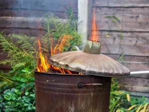 A metal bin ablaze with flames, surrounded by lush greenery. A metal brazier ablaze with flames, surrounded by lush greenery.